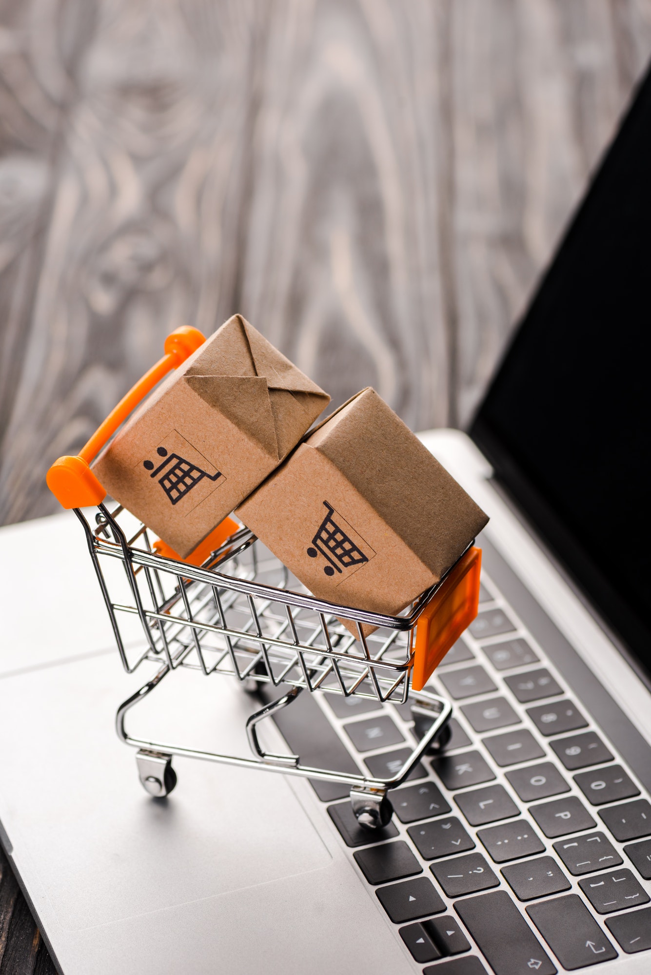 selective focus of toy carton boxes in small shopping cart on laptop with blank screen, e-commerce