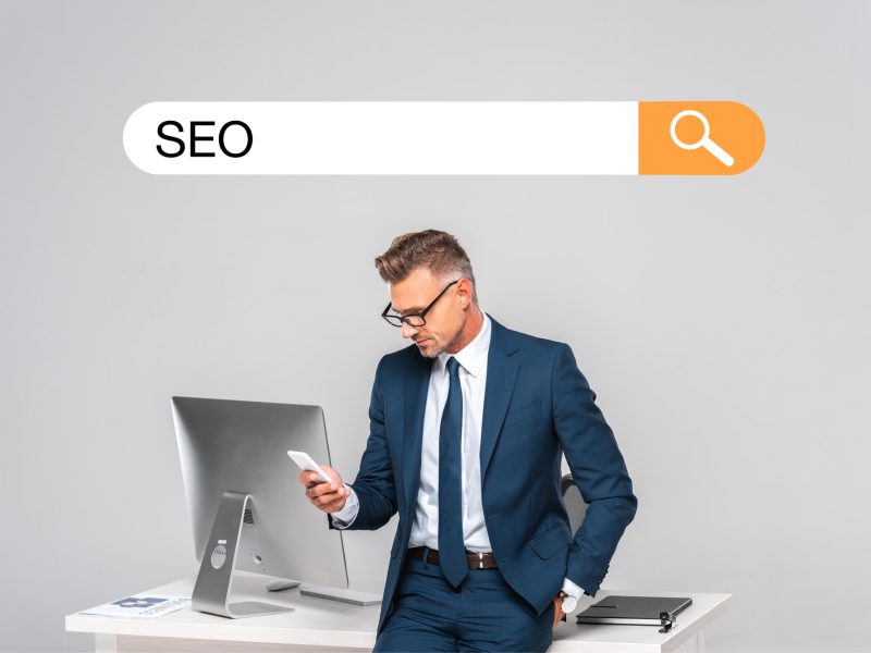 handsome businessman leaning on table and using smartphone isolated on grey with seo in search bar